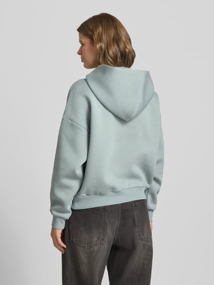 Vrouwenmodel draagt hoodie met logo en kangoeroezak van review, katoenmix, relaxed fit, logoprint, kangoeroezak. Ganzebody van achteren.