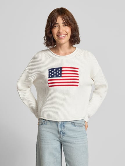 Frauenmodel trägt Strickpullover mit überschnittenen Schultern von GAP, reine Baumwolle, weiß, grob gestrickt, vorne USA-Flagge, Regular Fit, Rundhalsausschnitt, Ganzkörperansicht von vorne.