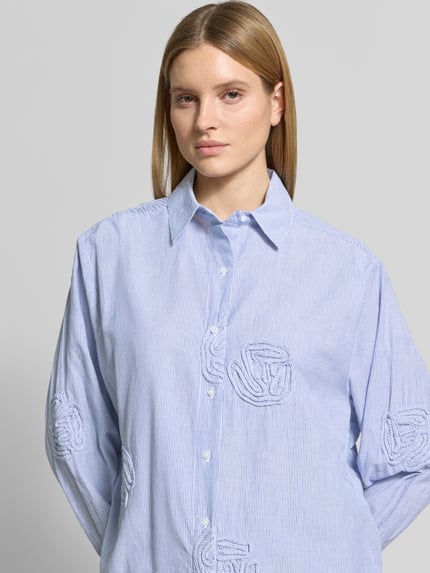 Modelka nosi Relaxed Fit bluzkę z wzorem w paski model 'Dalma Dori' od Neo Noir z czystej bawełny, kołnierzykiem koszulowym, florystycznymi aplikacjami, długość tyłu w rozmiarze 36: 73 cm, długość rękawów w rozmiarze 36: 60 cm, jasnoniebieskie i białe paski, faktura gładka, widok całego przodu, zbliżenie na górną część ciała.
