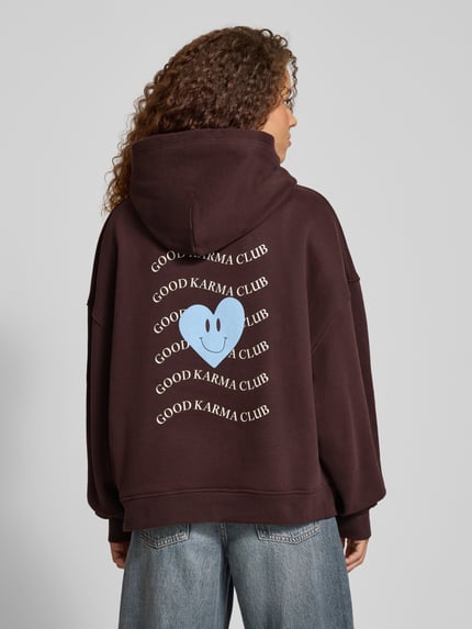 Frauenmodel trägt Hoodie mit rückseitigem Print Modell 'Good Karma Wave' von OH APRIL in Dunkelbraun, lockere Passform, Baumwoll-Mix, Ganzkörperansicht Rückansicht