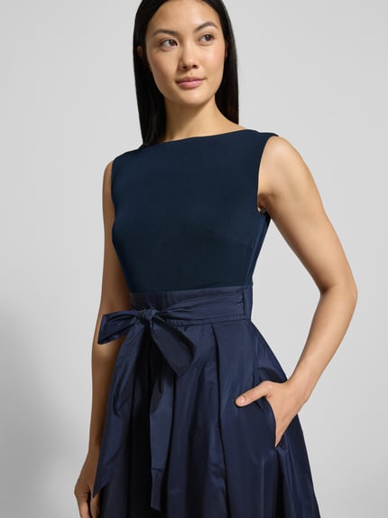 Frauenmodel trägt Abendkleid mit Bindegürtel und rückseitigem Reißverschluss von Christian Berg Cocktail, Material-Mix mit Stretch-Anteil, A-Linie, Midikleid, Rundhalsausschnitt, dunkelblau, glatte Textur, Nahaufnahme Oberkörper.