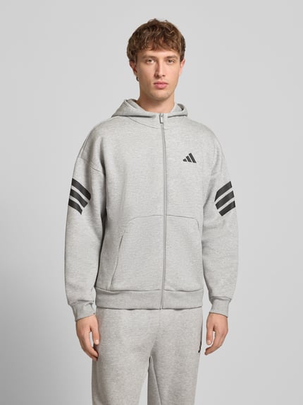 Männermodel trägt Sweatjacke mit Kapuze und Logo von adidas Sportswear, grau, Material-Mix mit Baumwoll-Anteil, Relaxed-Fit, Känguru-Tasche, Reißverschluss, Logo-Print, Ganzkörperansicht von vorne.