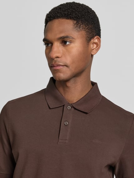 Męski model nosi Regular Fit Poloshirt aus reiner Baumwolle od s.Oliver RED LABEL, kolor brązowy, gładki, pikowana faktura, krótki rękaw, kołnierzyk polo, krótkie zapięcie na guziki, przód, zbliżenie na górną część ciała.