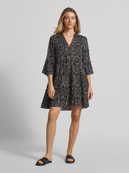 Frauenmodel trägt Ausgestelltes Blusenkleid aus reiner Viskose Modell 'EASY JOY' von Vero Moda, Optik:Allover-Muster, Mini-Länge, Henley-Ausschnitt, Raffung, 3/4-Arm mit Trompetenärmel, gerader Saum, Material: reine Viskose, schwarze Grundfarbe mit weißen Linien, glatte Textur, Ganzkörperansicht von vorne.