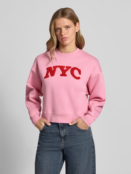 Frauenmodel trägt Relaxed Fit Sweatshirt aus Baumwoll-Mix Modell 'Tatum' von JJXX in Rosa mit rotem Statement-Patch, Rundhalsausschnitt, Rippbündchen und überschnittenen Schultern, Damen Sweatshirt, Ganzkörperansicht von vorne.