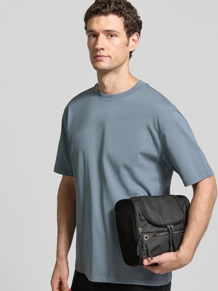 Männermodel trägt Kulturtasche mit Label-Details Modell 'Blanc Delight' von bugatti in Schwarz mit Reißverschluss, Nahaufnahme Oberkörper.