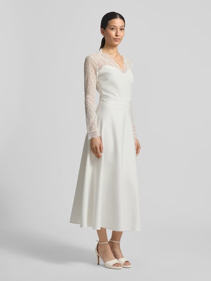 Frauenmodel trägt Tailliertes Brautkleid mit V-Ausschnitt Modell 'Joanna' von GUIDO MARIA KRETSCHMER WOMAN, Elastischer-Material-Mix, Oberteil aus Spitze, langärmelig, blickdichter Rockteil, Produkt in Creme, Ganzkörperansicht von vorne.