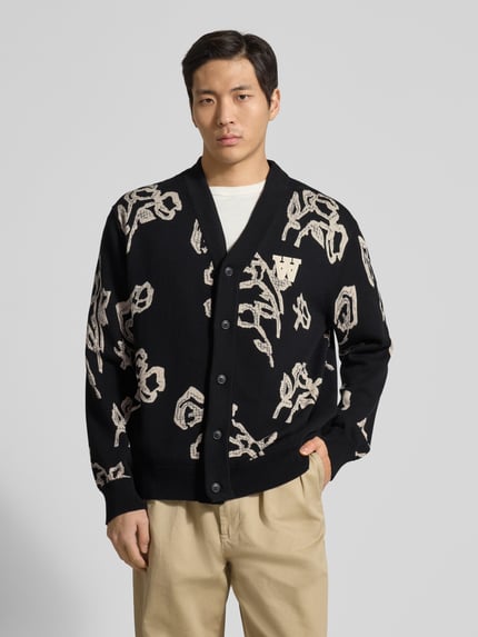 Männermodel trägt Cardigan im Allover-Look Modell 'Leif' von Wood Wood aus reiner Bio-Baumwolle, Regular-Fit, V-Ausschnitt, Knopfleiste, schwarze und beige Farben, mittlere Struktur, Nahaufnahme Oberkörper.