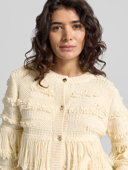 Frauenmodel trägt Strickjacke mit Fransen Modell 'AMELIE' von BRAX in unifarbenem Creme, strukturierte Strickoptik, Nahaufnahme Oberkörper.