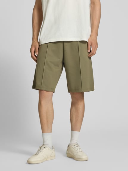 Männermodel trägt Shorts mit elastischem Bund und Ziernähten von REVIEW, Loose-Fit Material-Mix mit Stretch-Anteil, Bundweite 42 cm, Innenbeinlänge 26 cm, olivegrün, seitliche Eingrifftaschen, angedeutete Gesäßleistentaschen, Ganzkörperansicht von vorne.