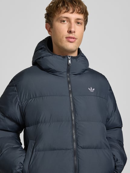 Männermodel trägt Regular Fit Puffer Jacket mit Kapuze von adidas Originals, jednokolorowy, krój regular fit, zamek błyskawiczny, detal z logo, długi rękaw, wykończenie rękawów i dołu elastyczne, hydrofobowy, kieszenie zapinane na zamek błyskawiczny, wykończenie materiału: tkanina, Nahaufnahme górnej części ciała.