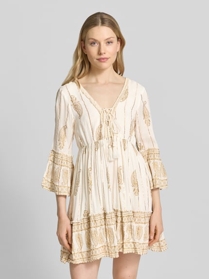 Frauenmodel trägt Minikleid mit Allover-Paisley-Print von Apricot aus Viskose-Mix, Taillierte Passform, V-Ausschnitt, 3/4-Arm, Schnürverschluss, Rüschenbesatz, in Creme und Beige-Tönen, Stoff mit leichter Struktur, Nahaufnahme Oberkörper.