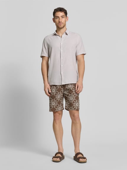 Männermodel trägt Shorts aus Leinen-Mix mit elastischem Bund und Kordelzug von MCNEAL, Relaxed-Fit, braunes Muster, Ganzkörperansicht von vorne. Herren Shorts aus Leinen-Baumwoll-Anteil mit seitlichen Eingrifftaschen und Gesäßtasche, Bundweite 40 cm, Innenbeinlänge 23 cm, Größe M.