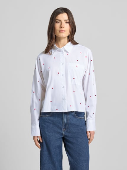 Frauenmodel trägt Boxy Fit Hemdbluse aus reiner Baumwolle von Tom Tailor Denim, Weiß mit roten Herzstickereien, Ganzkörperansicht von vorne.