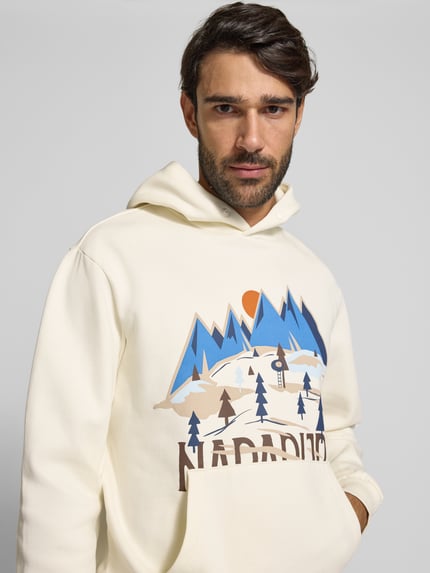Männermodel trägt Hoodie mit Label-Print von Napapijri in Creme mit blau-braunem Print, Baumwollmischung, Nahaufnahme Oberkörper. Rückenlänge Größe M: 64 cm, Ärmellänge: 73 cm, lockerer Schnitt, Känguru-Tasche, elastische Rippbündchen, Kapuze mit Knopf.