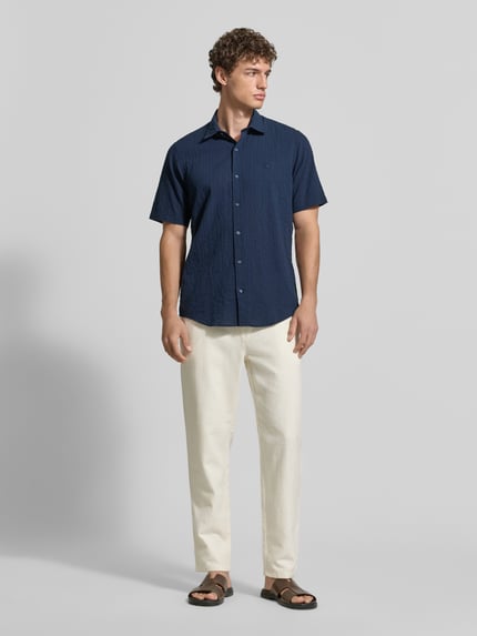 Męskimodel nosi Relaxed Fit Freizeithemd mit Logo-Stitching und Kentkragen von Lerros, seersucker, krój relaxed fit, kołnierzyk typu kent, listwa guzikowa, wyhaftowane logo, zaokrąglony dół, długość tyłu w rozmiarze M: 75 cm, szerokość w rozmiarze M: 45 cm, długość rękawów w rozmiarze M: 25 cm, ciemnoniebieski, faktura lekko pofalowana, gładkie jasne spodnie, sandały, całopostaciowy widok z przodu.