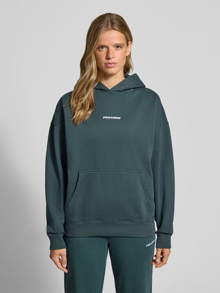 Frauenmodel trägt Oversized Fit Hoodie aus Baumwoll-Mix Modell 'CLARITA' von PEGADOR in Dunkelgrün mit Känguru-Tasche und Label-Detail, elastische Abschlüsse, Ganzkörperansicht von vorne.