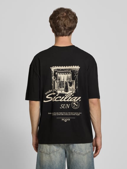 Männermodel trägt Oversized T-Shirt mit rückseitigem Print Modell 'Pedroche' von Pegador in Schwarz, Motiv in Beige, Puur katoen, oversized fit, Ganzkörperansicht Rückansicht.