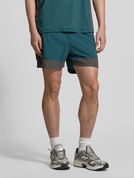 Męski model nosi szorty HALO DIVISION od HALO w kolorze zielonym z szarymi detalami, krój regular fit, elastyczny pas, kieszenie zapinane na zamek błyskawiczny, szerokość w rozmiarze M: 40 cm, długość wewnętrzna nogawki: 10 cm, całkowity widok z przodu