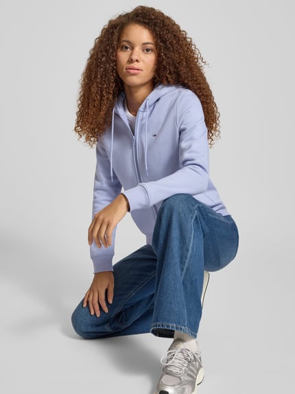 Modelka kobieca nosi Regular Fit Sweatjacke aus Baumwoll-Mix od Tommy Jeans, jasnoniebieska, długi rękaw, kaptur, zamek, widok z przodu w półzbliżeniu.