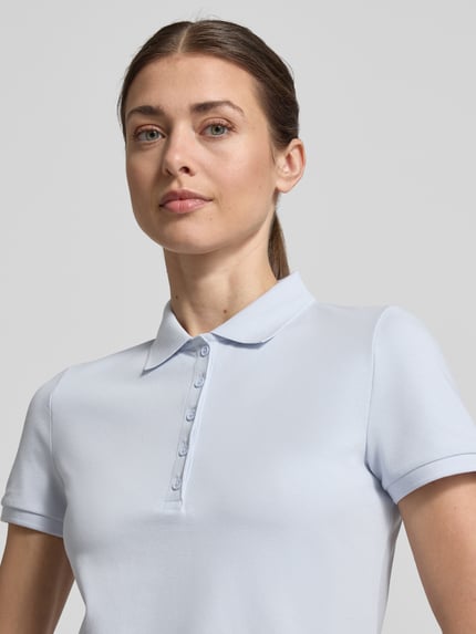 Modelka kobieca nosi Slim Fit Poloshirt mit Knopfleiste von Christian Berg Woman z mieszanki bawełny i elastanu, krój slim fit, listwa guzikowa, boczne rozcięcia, długość tyłu w rozmiarze S: 62 cm, jasny kolor, gładka tekstura, widok z przodu, zbliżenie na górną część ciała.