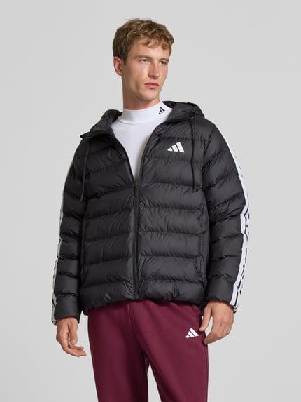 Männermodel trägt schwarze Regular Fit Steppjacke mit Kapuzenkragen von adidas Sportswear, weiße Logodetails, Ganzkörperansicht von vorne.