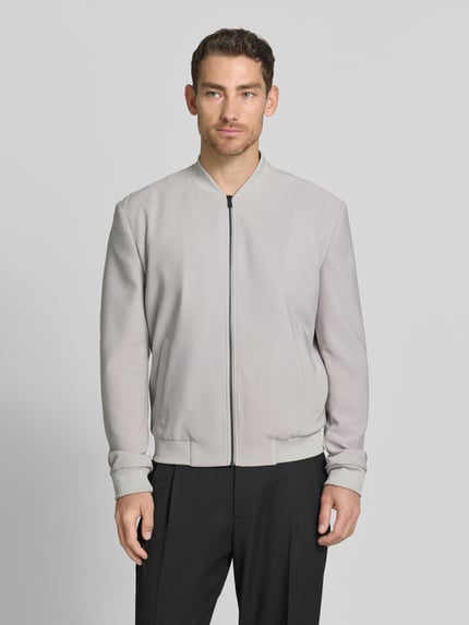 Męski model nosi bluzon z wpuszczanymi kieszeniami od MCNEAL, mieszanka materiałów z dodatkiem streczu, krój regular fit, stójka, zamek błyskawiczny, elastyczne wykończenia, długość tyłu w rozmiarze M: 65 cm, szerokość ramion w rozmiarze M: 45 cm, szerokość klatki piersiowej w rozmiarze M: 60 cm, długość rękawów w rozmiarze M: 71 cm. Jasnoszary, gładka struktura, proste formy. Ganzkörperansicht von vorne.