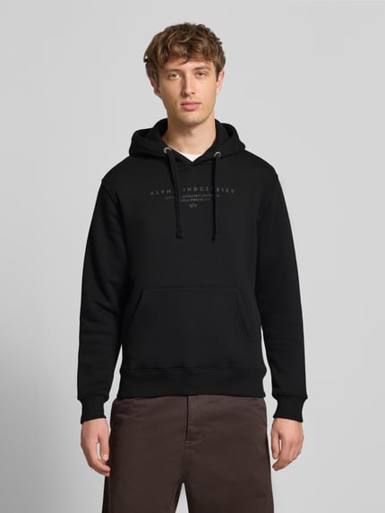Männermodel trägt Relaxed Fit Hoodie aus Baumwoll-Mix mit Label-Detail von Alpha Industries, Schwarz, Känguru-Tasche, Kapuze, Ganzkörperansicht von vorne.