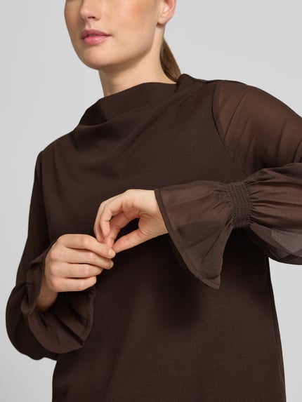 Frauenmodel trägt Relaxed Fit Langarmbluse mit Volants von s.Oliver BLACK LABEL in Dunkelbraun mit transparenten Ärmeln, Nahaufnahme Oberkörper.
