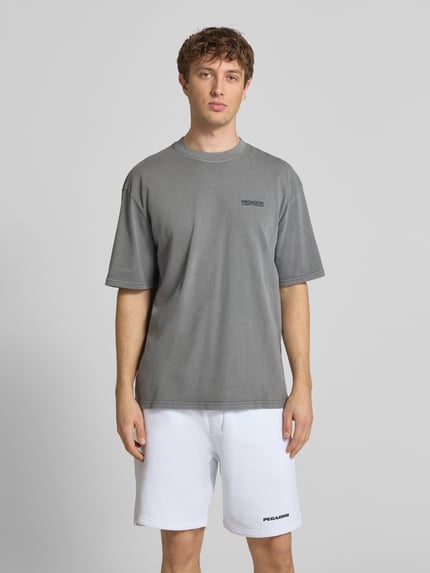 Męskimodel nosi Oversized T-Shirt aus Baumwolle Modell 'Hattem' von Pegador, kolor szary, detal z logo, okrągły dekolt, krój oversized fit, długość tyłu w rozmiarze M: 70 cm, przód, zdjęcie całej sylwetki
