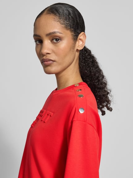 Vrouwenmodel draagt Sweatshirt mit Viskose-Anteil van monari in felrood, ronde hals met studs, statement-reliëf, elastische boorden, detailopname bovenlichaam.