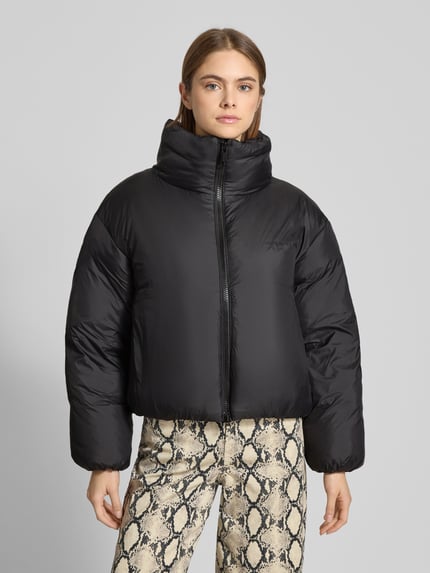 Kobiece modelka nosi czarną bomberkę Regular Fit Bomberjacke mit Zweiwege-Reißverschluss Modell 'Demi' von Carhartt Work In Progress, stójka, logo, długość tyłu 55 cm, szerokość ramion 54 cm, szerokość klatki piersiowej 58 cm, długość rękawów 54 cm, tekstura gładka, całkowity widok z przodu.