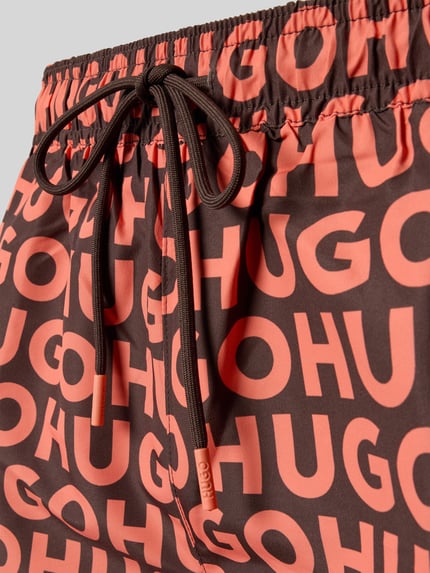 Produktfoto, Badehose mit Tunnelzug Modell 'KALY' von HUGO in Braun mit großem HUGO-Print und Tunnelzug, Textur glatt mit matter Oberfläche.
