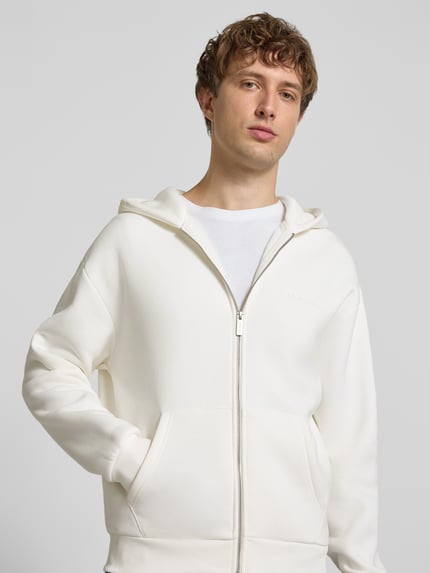 Männermodel trägt Regular Fit Sweatjacke mit Känguru-Tasche von REVIEW in Creme, Baumwoll-Mix, Reißverschluss, Kapuze, Nahaufnahme Oberkörper.