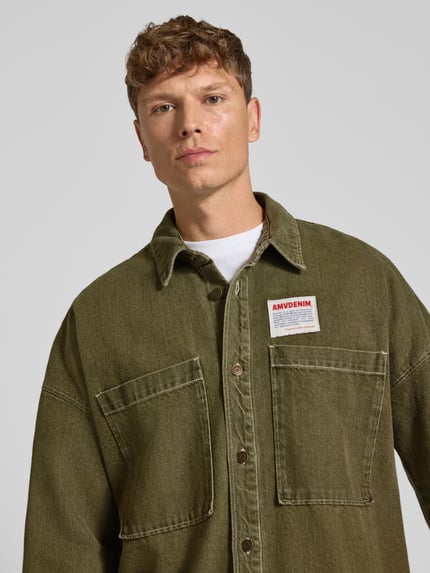 Männermodel trägt Relaxed Fit Jeanshemd mit Label-Detail Modell 'UZATOWN CHEMISES' von American Vintage in Olivgrün, Nahaufnahme Oberkörper. Reine Baumwolle, große Brusttaschen, Kentkragen, sichtbares Label auf der Vorderseite, strukturierte Stofftextur.