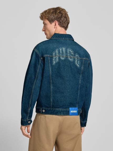 Mężczyznę model nosi kurtka jeansowa Regular Fit Jeansjacke aus reiner Baumwolle Modell 'Dex' von HUGO, niebieski dżins, beżowe spodnie, Ganzkörperansicht Rückansicht, kurtka HUGO Blue.