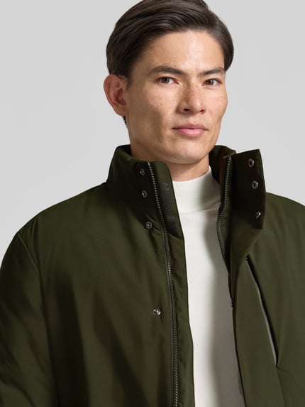 Männermodel trägt olivgrünen Mantel mit Kapuze und Brusttasche Modell 'SESLE' von Save The Duck, glatte Oberfläche, Nahaufnahme Oberkörper.