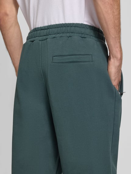 Männermodel trägt Loose Fit Sweatpants aus Baumwoll-Mix mit Label-Detail von PEGADOR in Dunkelgrün, elastischer Bund, Eingrifftaschen, Detailaufnahme Rückansicht.