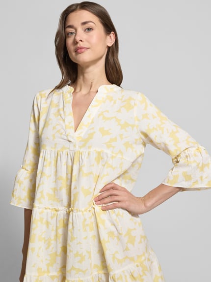 Frauenmodel trägt Ausgestelltes Blusenkleid aus reiner Baumwolle Modell 'DICTHE' von Vero Moda mit gelbem Allover-Muster, Henley-Ausschnitt und Trompetenärmeln, Mini-Länge, lockere Passform, Nahaufnahme Oberkörper.