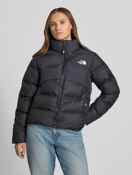 Modelka nosi czarną kurtkę pikowaną z wyhaftowanym logo, zamkiem błyskawicznym i kieszeniami zapinanymi na zamek błyskawiczny, Model 'SAIKURU' od The North Face, krój regular fit, stójka, widok całej sylwetki z przodu, długość tyłu w rozmiarze S: 59 cm, szerokość ramion w rozmiarze S: 43 cm, szerokość w klatce piersiowej w rozmiarze S: 53 cm, długość rękawów w rozmiarze S: 62 cm.