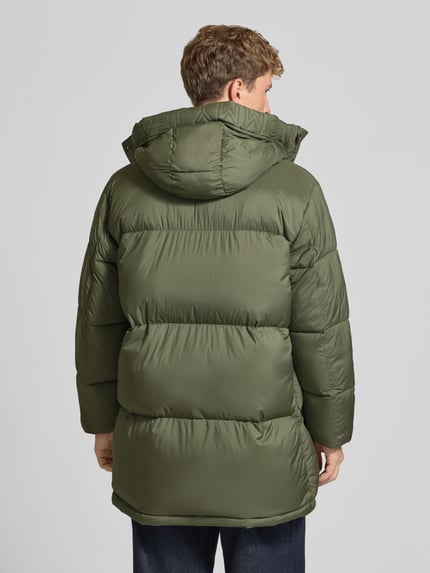 Männermodel trägt Parka mit Logo-Patch Modell 'WORKWEAR' von Tommy Jeans in matt-olivgrün, Struktur, Longline, Ganzkörperansicht Rückansicht.