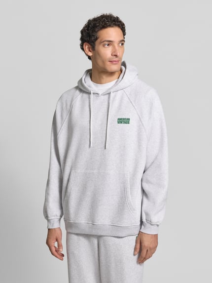 Männermodel trägt Sweatshirt mit Kapuze und Kängurutasche von AMERICAN VINTAGE in hellgrauer Melange-Optik mit Logo-Schriftzug, Ganzkörperansicht von vorne.