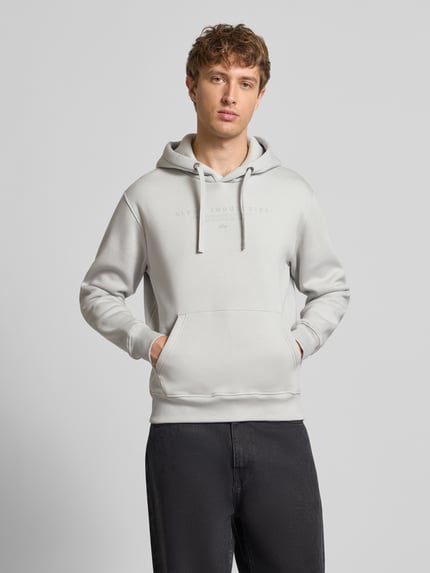 Männermodel trägt Relaxed Fit Hoodie aus Baumwoll-Mix mit Label-Detail von Alpha Industries in Hellgrau mit Känguru-Tasche, Ganzkörperansicht von vorne.