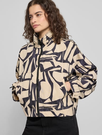 Frauenmodel trägt Oversized Hemdjacke mit Allover-Muster Modell 'BETTY BATSLEEVE' von Only in Beige-Schwarz, grafisches Muster, elastische Abschlüsse, Stehkragen mit Kapuze, Reißverschluss, seitliche Taschen, cropped Länge, Ganzkörperansicht von vorne.
