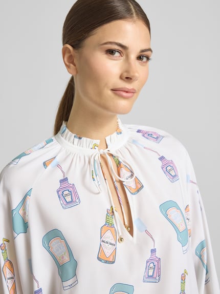 Vrouwenmodel draagt Regular Fit Cardigan aus Seiden-Mix in Strick-Optik van HERZENS, wit met kleurrijke illustraties, elastische seiden-mix, getailleerde pasvorm, afneembare stoffen riem, ruglengte 62 cm, mouwlengte 55 cm, close-up bovenlichaam.