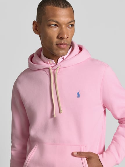 Männermodel trägt Regular Fit Hoodie aus Baumwoll-Mix von Polo Ralph Lauren in Rosa mit blauer Logo-Stickerei, Nahaufnahme Oberkörper.