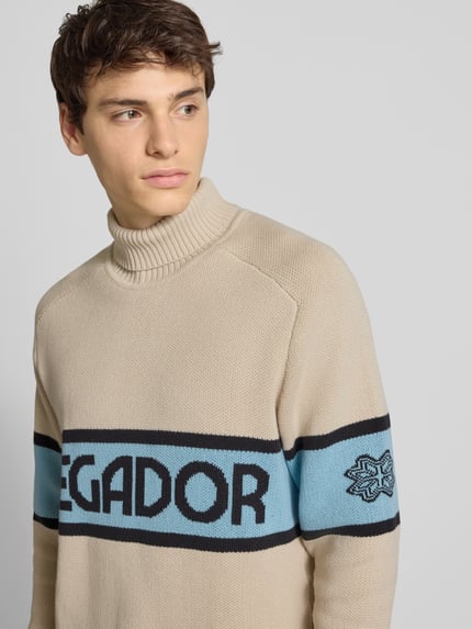 Männermodel draagt Strickpullover mit Rollkragen van Pegador in beige met blauwe en zwarte details, katoenmix, labelprint, Nahaufnahme bovenlichaam.