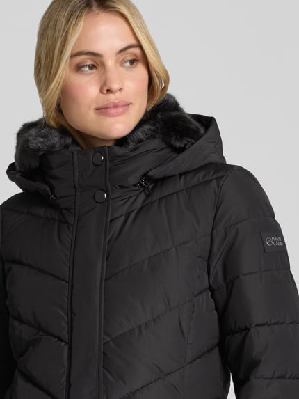 Frauenmodel trägt schwarze Jacke mit abnehmbarer Kapuze von NAUTGÅRD, gesteppt, sichtbarer Label-Patch, Nahaufnahme Oberkörper.