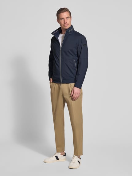 Männermodel trägt Regular Fit Jacke mit Stehkragen von bugatti in Dunkelblau mit Logo-Detail, verstaubarer Kapuze, Zweiwege-Reißverschluss, Eingrifftaschen, elastischen Ärmelabschlüssen, helle Hose, Sneaker, Ganzkörperansicht von vorne. Rückenlänge 68 cm, Schulterbreite 42 cm, Brustweite 50 cm, Ärmellänge 65 cm bei Größe 50.