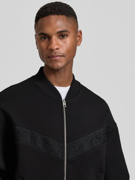 Männermodel trägt Oversized Sweatjacke aus Baumwoll-Mix Modell 'DROMZIP' von HUGO in Schwarz, Logo-Patch, Nahaufnahme Oberkörper.